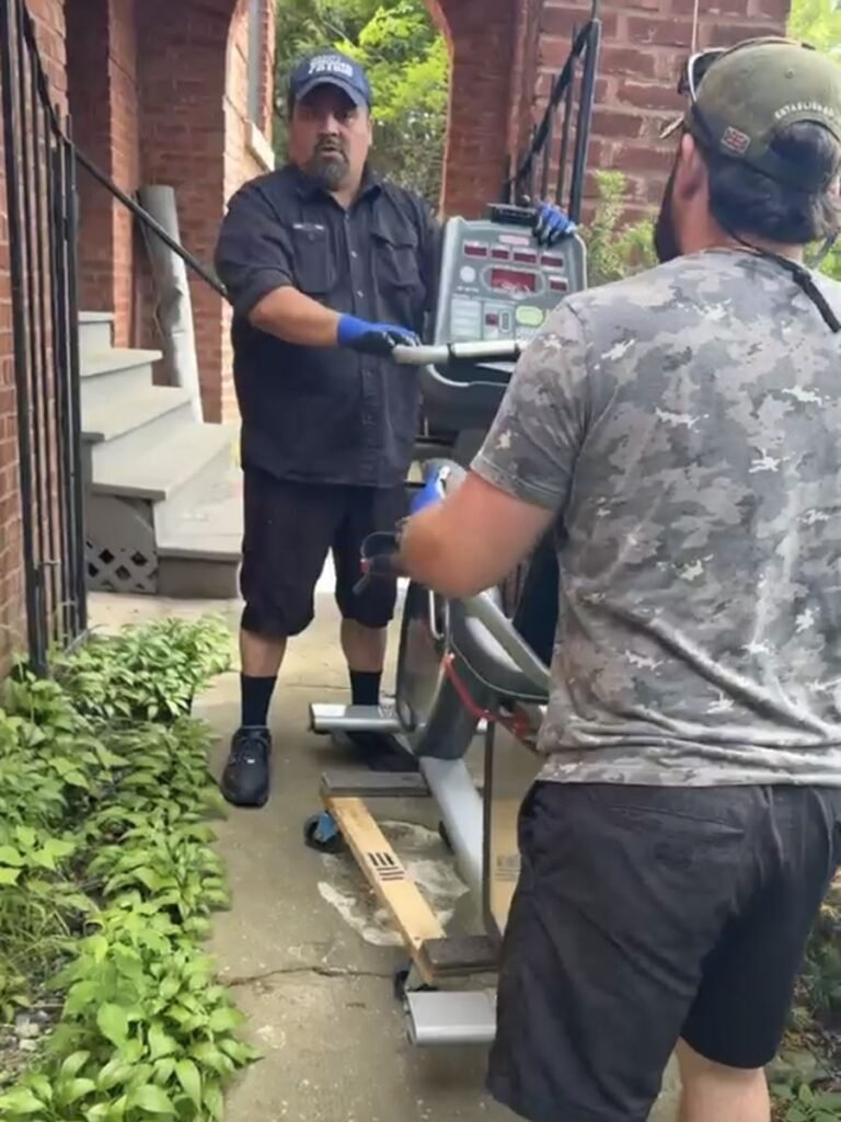 Two crew members carrying an elliptical machine out of a house