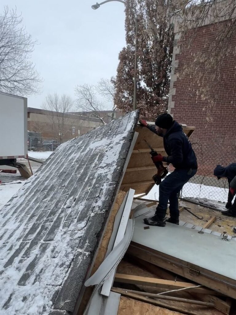 Crew member taking apart the shingled roof of a shed in winter
